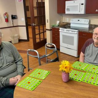 Two people sitting at a table in a kitchen area with bingo cards and a vase of flowers.