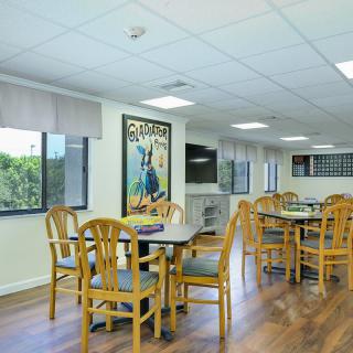 A bright community dining area with several tables and chairs, featuring large windows.