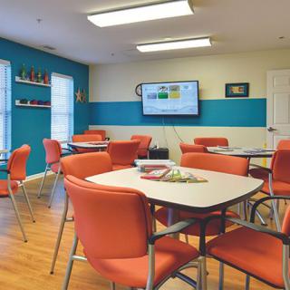 A modern classroom with orange chairs and tables, featuring a television on the wall.