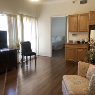 A bright living space with hardwood floors, featuring a TV, chairs, and kitchen cabinets.
