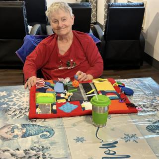 An elderly person sitting at a table with an activity board, smiling contentedly.