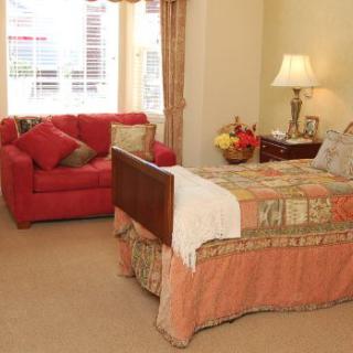 A cozy bedroom featuring a bed with a colorful quilt, a red sofa, and a lamp on a bedside table.
