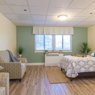 A cozy bedroom with a comfortable bed, an armchair, and decorative plants, featuring a large window that brightens the space.