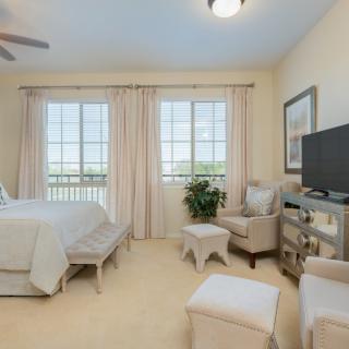 A well-lit bedroom with neutral tones, featuring a bed, armchair, television, and large windows with curtains.