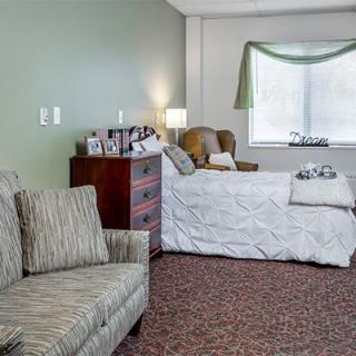 A cozy bedroom featuring a bed, armchair, and dresser with soft lighting.