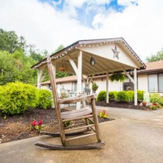 A rustic wooden rocking chair sits on a porch in front of a building with a landscaped garden and covered entrance.
