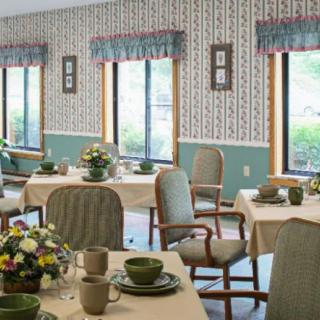 A cozy dining room with elegantly set tables and chairs, adorned with floral arrangements and large windows providing natural light.