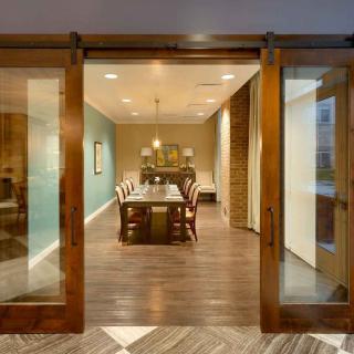 A modern dining room with wooden flooring, featuring a long table and chairs, illuminated by a chandelier and accessed through sliding glass doors.