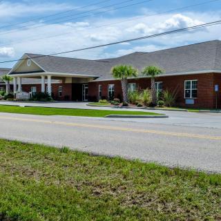 A one-story brick building with a covered entrance, situated alongside a road with palm trees and grassy surroundings under a partly cloudy sky.