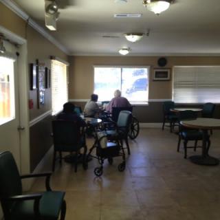 A calm nursing home common area with seniors sitting at tables near a window.