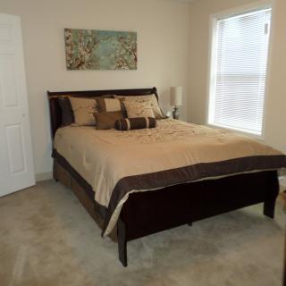 A cozy bedroom with a neatly made bed, soft carpeting, and wall artwork next to a large window.