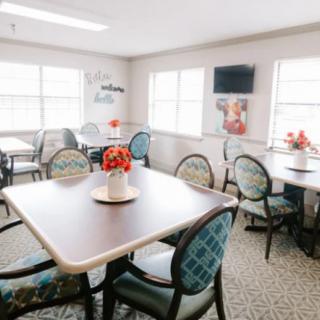 A bright dining room with multiple tables and chairs, decorated with flowers and featuring a wall-mounted television.