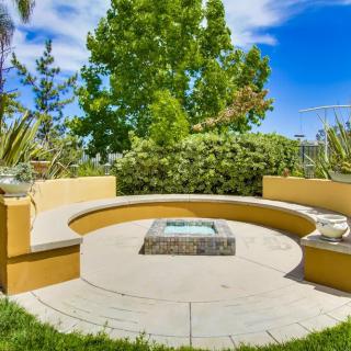 A sunny outdoor patio with a circular seating area and a central fire pit surrounded by lush greenery.
