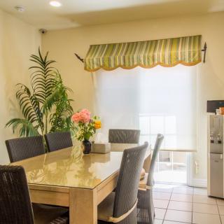 A cozy dining room with a table set, surrounded by chairs and decorated with a flower vase, plant, and window curtains.