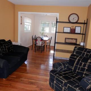 A cozy living room with a sofa set and an adjacent dining area, featuring wooden flooring and a shelving unit.