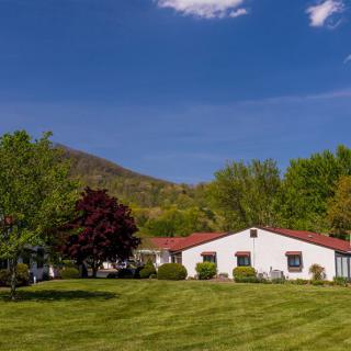 A picturesque house with a red roof surrounded by lush green grass and trees, set against a backdrop of mountains under a clear blue sky.