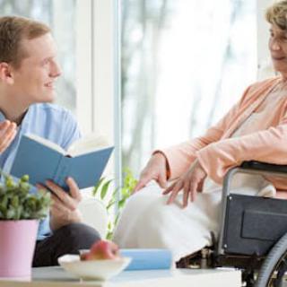 A caregiver reads to a person in a wheelchair inside a bright room.