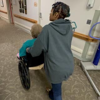 A caregiver pushing an elderly person in a wheelchair down a hospital corridor.