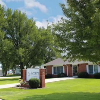 A quaint brick house surrounded by trees with a neatly manicured lawn and a sign in front.