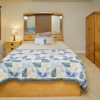 A cozy bedroom with a neatly made bed featuring a blue and white quilt, surrounded by wooden furniture and a glowing lamp on the nightstand.