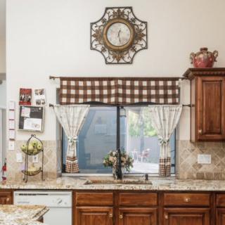 A cozy kitchen interior featuring wooden cabinets, a decorative clock, and checkered curtains over the window.