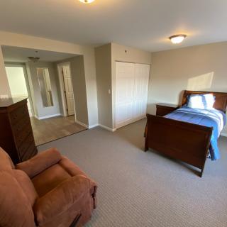 A spacious bedroom with a single bed, armchair, and dresser, featuring a carpeted floor and ample natural light.