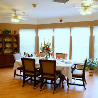 A well-lit dining room with a table set for six, featuring a floral centerpiece and surrounded by large windows.