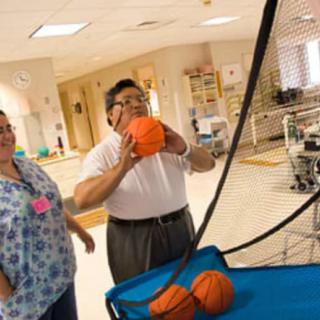 An elderly man participates in a basketball activity as part of a rehabilitation or therapy program, assisted by a caregiver.