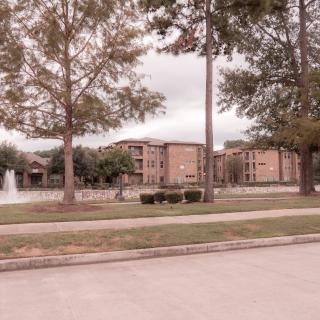 A view of an apartment complex with a fountain surrounded by trees on a cloudy day.
