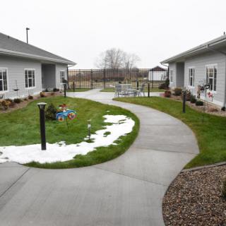 A serene pathway connecting two buildings, framed by patches of grass and remnants of snow under a gray sky.