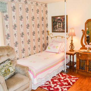 A cozy bedroom with vintage decor, featuring a single bed, armchair, and ornate mirror.