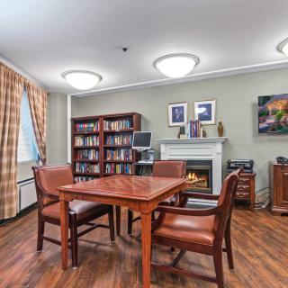 A cozy library room with a wooden table and chairs, surrounded by bookshelves and decorated with artwork above a fireplace.