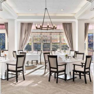 An elegant dining room with neatly arranged tables and chairs, complemented by a chandelier and large windows with curtains.