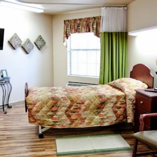 A cozy hospital room featuring a neatly made bed, green curtains, and wooden flooring.