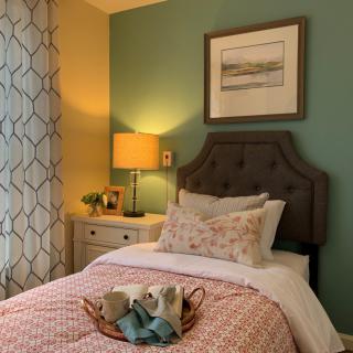A cozy bedroom with a neatly made bed, adorned with decorative pillows and a comforter, accompanied by a bedside lamp and artwork on the wall.