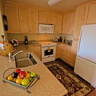 A modern kitchen with wooden cabinets, featuring a fridge, oven, microwave, and a fruit basket on the countertop.