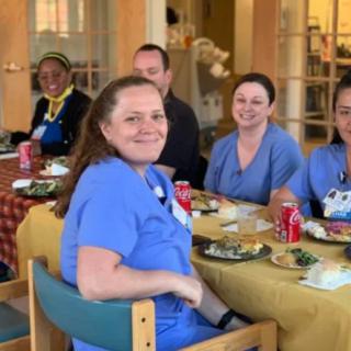 A group of people wearing scrubs enjoying a meal together indoors, with smiles on their faces.