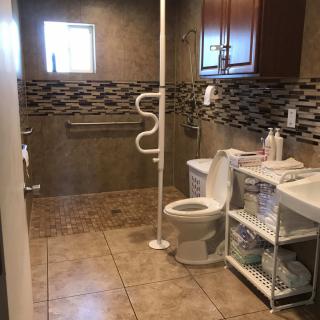 A spacious and accessible bathroom with tiled walls and floor, featuring a toilet, sink, shower area, and grab bars for support.