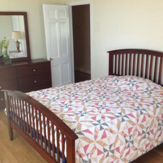 A tidy bedroom with a wooden bed frame and colorful quilt, complemented by a dresser with a mirror.