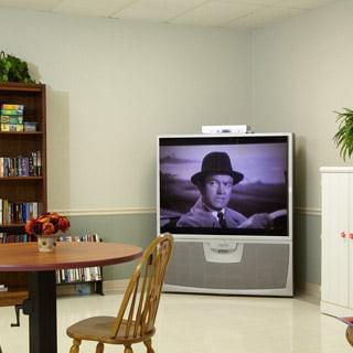 A cozy living room with a television displaying a black and white movie, a bookshelf, and a wooden table with chairs.