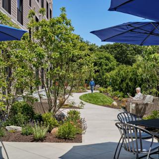 A peaceful outdoor patio area with tables, chairs, and blue umbrellas surrounded by greenery and a garden pathway.
