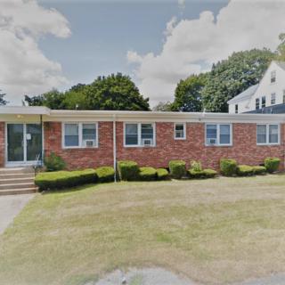 A single-story brick house with a neatly trimmed lawn and shrubs, set in a residential neighborhood under a partly cloudy sky.