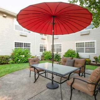 A patio with a glass table and chairs, shaded by a large red umbrella in a garden setting.