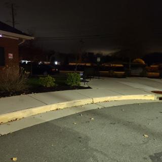 A quiet evening scene showing a sidewalk and parking area under dim lighting.