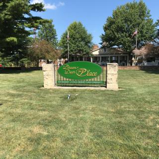 A welcoming green sign on a neatly maintained lawn with trees and a building in the background.