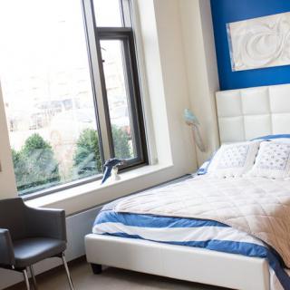 A modern bedroom with a blue accent wall, featuring a bed, chair, and large window.