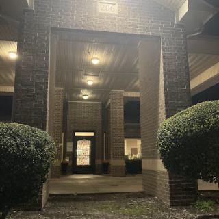 A well-lit entrance to a building at night, framed by neatly trimmed bushes.