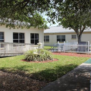 A modest building with a small garden, white fence, and surrounding trees.