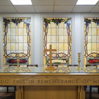 A church altar with a communion table adorned with a cross and candles, set against stained glass windows.