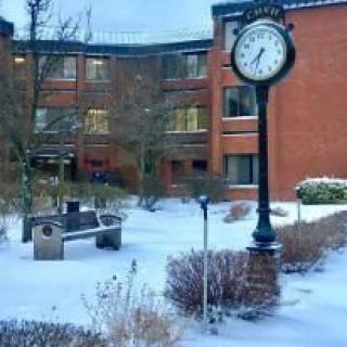 A snowy courtyard with a clock and benches in front of a brick building.